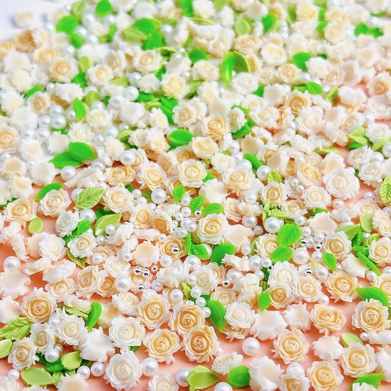 May include: A collection of small, white and cream-colored flower embellishments, perfect for crafting and decorating. The embellishments are scattered on a light pink surface, along with small white pearls.
