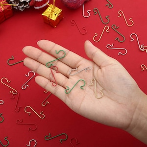 May include: A selection of colourful metal ornament hooks in red, green, silver, and gold. The hooks are scattered on a red surface, with some held in a hand. The hooks are in a variety of colours, perfect for Christmas tree decorations.