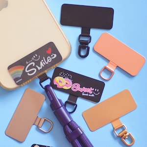 May include: A set of five phone charms with different designs. The charms are black, gold, and pink. The charms have a ring on the top for attaching to a phone case. The charms feature the words "Smiles", "Sweet", and a donut design.