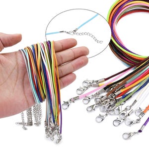 Puede incluir: Un juego de 20 cordones de algodón encerado de colores vivos con cierres de langosta plateados. Los cordones están dispuestos en una mano, y algunos están dispuestos sobre una superficie blanca.