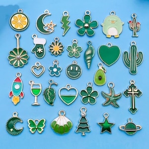 May include: A selection of green and gold enamel charms in diverse shapes, such as a crescent moon, flower, cactus, and rocket. The charms are set against a light blue backdrop. These charms are perfect for jewellery making.