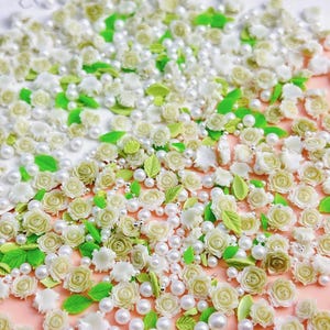 May include: A collection of white and green floral embellishments, including small roses, leaves, and pearls. These delicate decorations are perfect for adding a touch of elegance to crafts, nail art, or other projects.