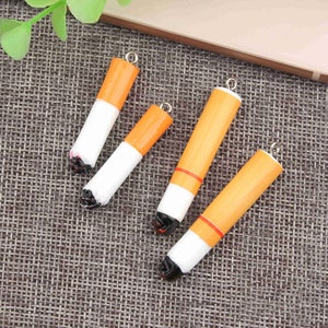 May include: Four miniature cigarette charms with a gold-colored loop at the top. The charms are made of white and orange resin and have a black painted tip.