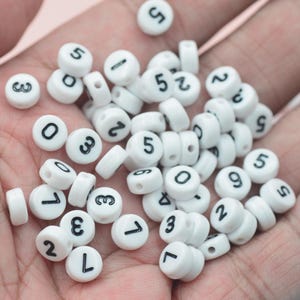 May include: A collection of white plastic round beads with black numbers from 0 to 9. The beads are scattered in a hand.
