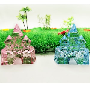 May include: Two miniature crystal castle figurines, one pink and one blue, on a green grass background.