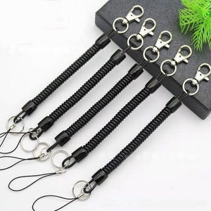 May include: Five black coiled keychains with silver-colored metal clasps and key rings. Each keychain has a black cord loop attached to the key ring. The keychains are arranged diagonally across a white surface.