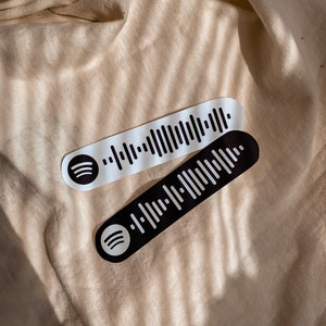 May include: Two black and white Spotify barcode stickers with the Spotify logo. The stickers are on a beige fabric background.