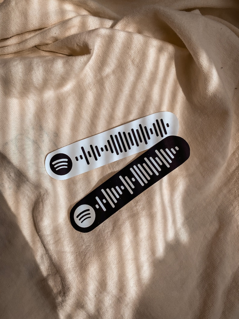 May include: Two black and white Spotify barcode stickers with the Spotify logo. The stickers are on a beige fabric background.
