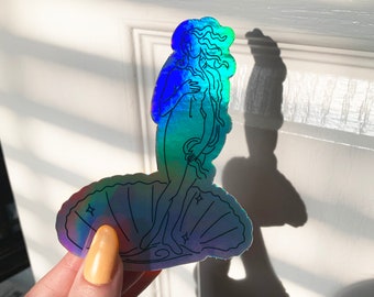 May include: A holographic sticker featuring a line drawing of the Birth of Venus, a famous Renaissance painting. The sticker is iridescent and has a blue and green color scheme.