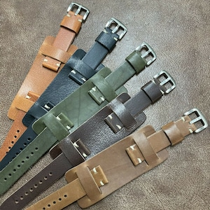 Op de afbeelding: Vijf verschillende leren horlogebandjes in verschillende kleuren, waaronder bruin, groen, zwart en beige. De bandjes hebben witte stiksels en zilveren gespen.