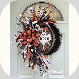 Puede incluir: Una corona de Halloween con base de ramas negras, un letrero ovalado con un fantasma y el texto BOO, y un gran lazo con estampado naranja y negro. El arreglo incluye follaje blanco y negro, bayas naranjas y detalles en color blanco.
