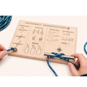 Peut inclure: Un panneau de nœuds en bois avec des schémas et une barre métallique pour pratiquer les nœuds nautiques. Le panneau porte le texte "KNOTENBRETT - SPORTBOOTSSCHEIN". Une corde bleue et jaune est utilisée pour démontrer les nœuds. Le panneau est tenu par deux mains.