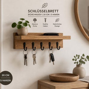 Puede incluir: Un llavero de madera de roble de 30 cm de ancho con un estante y cinco ganchos negros. Una pequeña maceta con una planta y llaves cuelgan de los ganchos. El texto en el llavero dice "SCHLÜSSELBRETT".