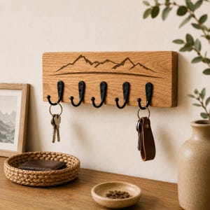 May include: A rectangular light brown wooden wall-mounted key holder featuring a laser-engraved mountain range design. It includes five black metal hooks, with two sets of keys hanging from the outer hooks on the left and right sides.
