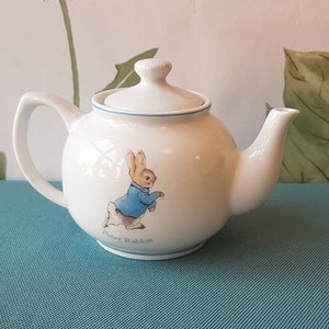 Op de afbeelding: Witte theepot van keramiek met een blauwe rand en een Peter Rabbit-illustratie. De theepot heeft een deksel en een tuit.