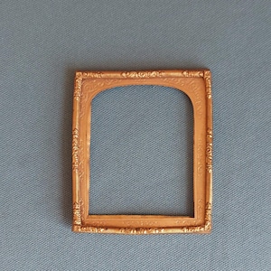 May include: Ornate gold-colored picture frame with a square shape and an arched top. The frame has detailed decorative carvings. The background is a textured, light blue fabric.