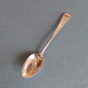May include: A silver spoon with a rounded bowl and a long handle. The spoon has a reflective surface and appears to be made of metal. The handle has engraved initials. The spoon is set against a grey background.