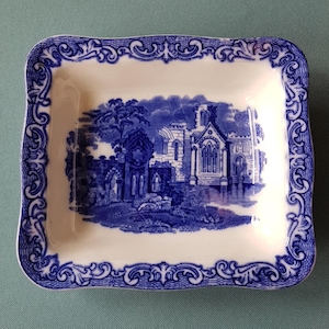 Peut inclure: Un plat en céramique blanche avec un motif bleu et blanc. Le motif représente un bâtiment avec un toit pointu et des arbres. Le plat a un bord festonné.