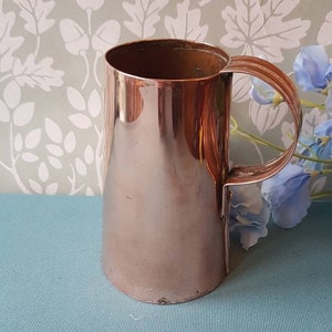 May include: A shiny copper mug with a handle. The mug has a rounded bottom and a slightly flared top.