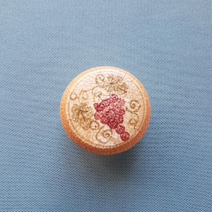 May include: A round cork bottle stopper with a decorative design. The design features a cluster of red grapes and gold-colored leaves. The stopper is set against a light blue background.