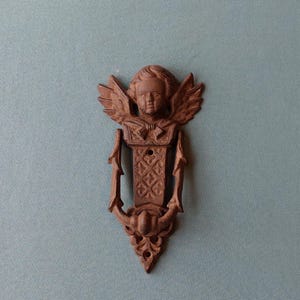 May include: A cast iron door knocker with an angel design. The knocker features a detailed face with wings and a decorative pattern on the body. The colour is a rustic brown, and the knocker is mounted on a light blue background.