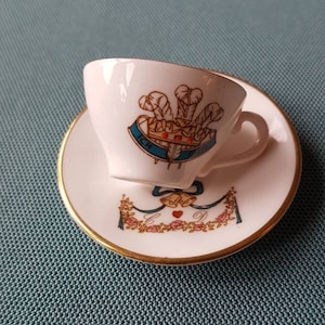 Puede incluir: Un juego de taza y platillo blanco con ribete dorado. La taza y el platillo presentan un diseño de cresta colorido con una corona, plumas y el texto "ICH". El platillo también tiene detalles florales y de cinta. Un juego de té coleccionable clásico.