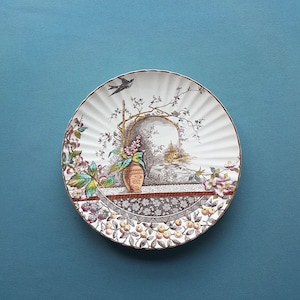 May include: A white ceramic plate with a scalloped edge features a colourful floral design with a bird, a vase, and a landscape scene. The plate has a gold rim.