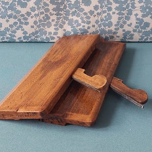 May include: A wooden book stand with a hinged design. The stand is made of a warm-toned wood and features a support arm to hold books at an angle. The background is a floral pattern on a light blue surface.