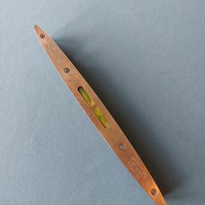 Puede incluir: Un nivel de latón antiguo con forma rectangular y extremos redondeados. El nivel tiene dos pequeños viales de vidrio llenos de un líquido amarillo-verdoso. El texto "Warranted" y "Made in England" es visible.