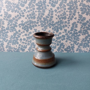 May include: A small, ceramic candle holder with a blue and brown glaze. The holder has a rounded base, a central section with a brown band, and a flared top. The background features a blue floral pattern.