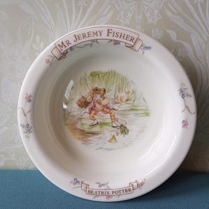 May include: A white ceramic bowl with a blue and white floral border. The centre of the bowl features a picture of a frog holding a fishing rod and a basket. The bowl has a banner that reads "Mr. Jeremy Fisher" and another banner that reads "Beatrix Potter".