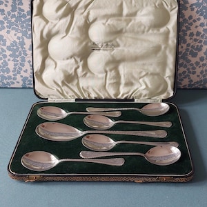 May include: A set of silver spoons displayed in an open, vintage case. The spoons are arranged on a green velvet surface, with the case's interior lined in a light green satin. The case is black with gold trim.