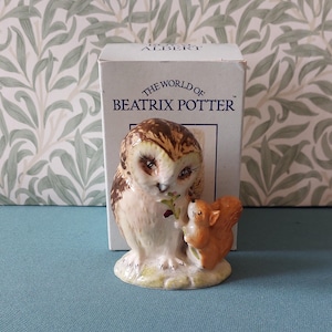 Könnte beinhalten: Keramikfigur einer Eule und eines Eichhörnchens aus der Sammlung "The World of Beatrix Potter". Die Eule ist braun und weiß, hält einen Zweig, und das Eichhörnchen ist orange. Die Figur steht auf einem hellgrünen Sockel, mit der Box im Hintergrund.