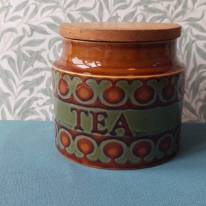 May include: A cylindrical ceramic tea canister with a glossy brown glaze and a wide green band featuring the word TEA in dark lettering. The container is decorated with a repeating scalloped pattern in brown and green and includes a flat wooden lid.