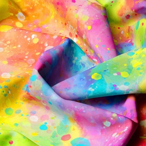 Op de afbeelding: Een levendige stof met een aquareleffect. Het ontwerp toont een mix van heldere kleuren, waaronder roze, geel, blauw en oranje, met witte verfspatten. De stof is gevouwen, waardoor schaduwen en diepte ontstaan.