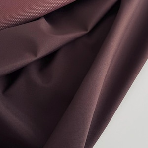 Pode incluir: Um tecido de cor ameixa profunda com um acabamento liso e mate, drapeado em dobras suaves. A secção superior do material apresenta um padrão de superfície texturado de grão fino que contrasta com o tecido liso e sólido por baixo.