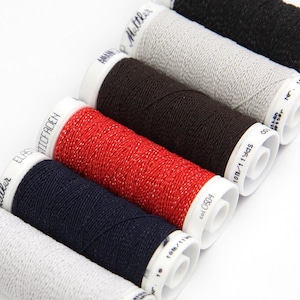 May include: Six spools of thread in various colors, including white, black, red, and navy blue. The spools are labeled with the brand name "Mettler" and the text "ELASTIC THREAD".