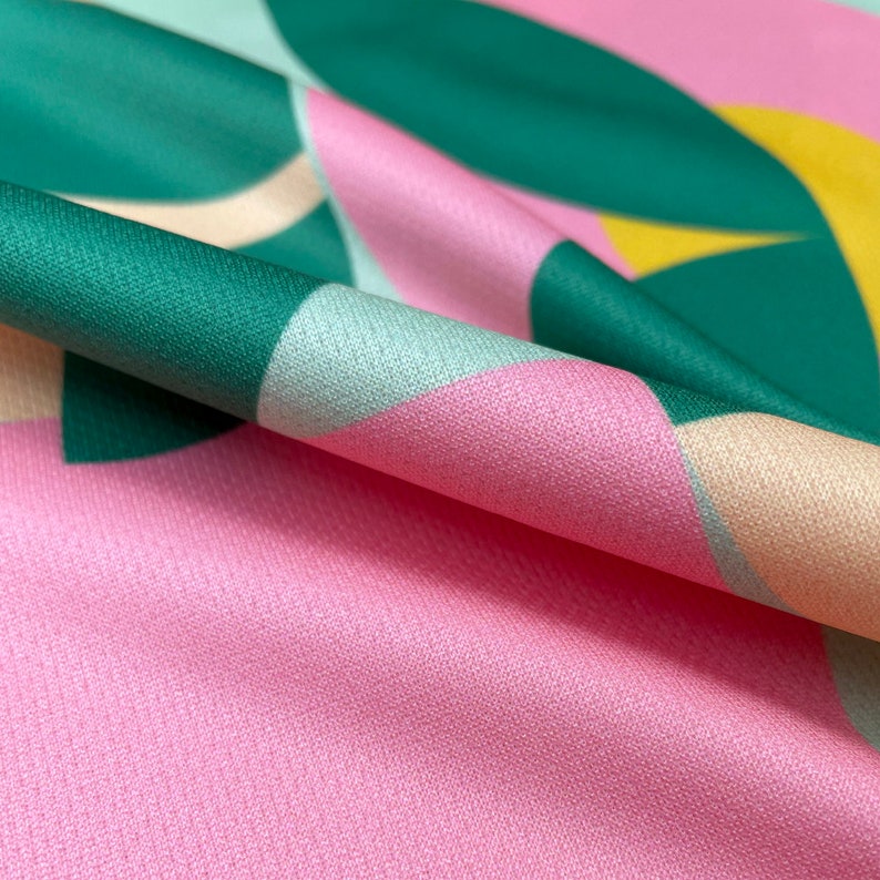 K&ouml;nnte beinhalten: Nahaufnahme eines rosa Stoffes mit einem geometrischen Muster aus gr&uuml;nen, blauen und gelben Formen. Der Stoff ist gefaltet und zeigt die Textur des Materials.