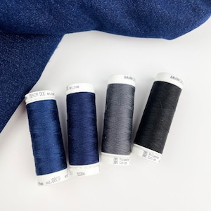 Pode incluir: Quatro carretéis de linha em tons de azul, cinza e preto são dispostos sobre uma superfície branca, com um tecido de ganga no fundo. Os carretéis têm rótulos brancos com texto, incluindo "DENIM DOC" e "78% Polyester 38% Cotton".