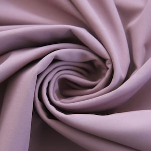 Op de afbeelding: Een effen mauvekleurige stof met een gladde, matte textuur, gerangschikt in een reeks zachte, vloeiende plooien en diepe, wervelende draperieën die een dynamisch patroon van licht en schaduw op het oppervlak van het textiel creëren.
