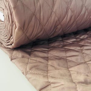 Peut inclure: Gros plan sur un tissu matelassé enroulé, de couleur rose pâle. Le tissu présente un motif en losanges créé par des lignes cousues. La matière semble douce et a un léger éclat, suggérant une finition satinée ou similaire. La texture est lisse et attrayante.