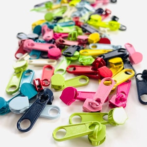 May include: A collection of colorful zipper pulls in various shades, including pink, green, yellow, and blue. The pulls are made of plastic and are scattered on a white surface. The image showcases a variety of shapes and sizes, suitable for sewing and craft projects.