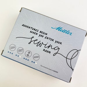 May include: A light blue box with the text "ADVENTURES BEGIN WHEN YOU ENTER YOUR sewing ROOM." The box has the brand name "Mettler" in blue and the text "The consumer brand of AMANN."