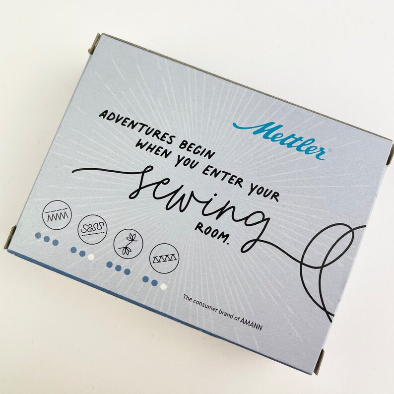 May include: A light blue box with the text "ADVENTURES BEGIN WHEN YOU ENTER YOUR sewing ROOM." The box has the brand name "Mettler" in blue and the text "The consumer brand of AMANN."