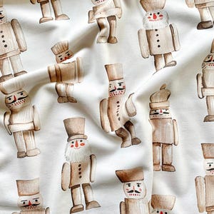May include: A white fabric with a repeating pattern of brown nutcracker figurines. The nutcrackers are all wearing different hats and have different facial expressions.