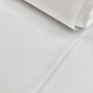 May include: A close-up shot of a roll of white, non-woven fabric. The fabric has a textured, mesh-like appearance. The roll is partially unraveled, revealing the layers of the material. The background is a plain white surface.