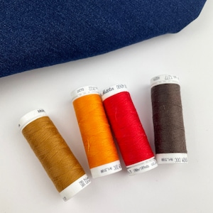 Pode incluir: Quatro bobinas de linha em tons de castanho, laranja, vermelho e castanho escuro estão dispostas sobre uma superfície branca, com um pedaço de tecido de ganga azul no fundo. As bobinas de linha estão rotuladas com texto. Adequado para costura e artesanato.