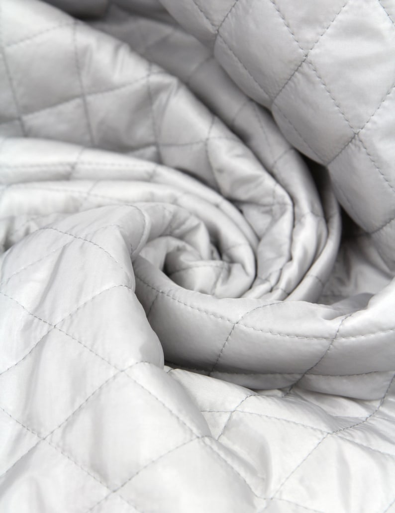 May include: Close-up of a white quilted fabric with a diamond pattern. The fabric is folded and twisted, creating a soft and textured look.