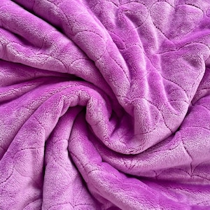 May include: Close-up of a soft, lavender blanket with a quilted leaf pattern. The fabric is plush and fluffy, with folds and shadows creating depth. The blanket's texture is prominent, giving a cosy feel. The colour is a vibrant shade of purple, adding to its appeal.