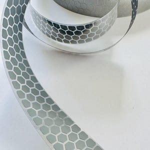 Könnte beinhalten: Eine Rolle silbernes Reflexband mit einem sich wiederholenden hexagonalen Wabenmuster. Das Band ist aufgerollt, wobei ein Ende nach außen ragt, um die metallische, gitterartige Textur der Oberfläche deutlich vor einem weißen Hintergrund zu zeigen.